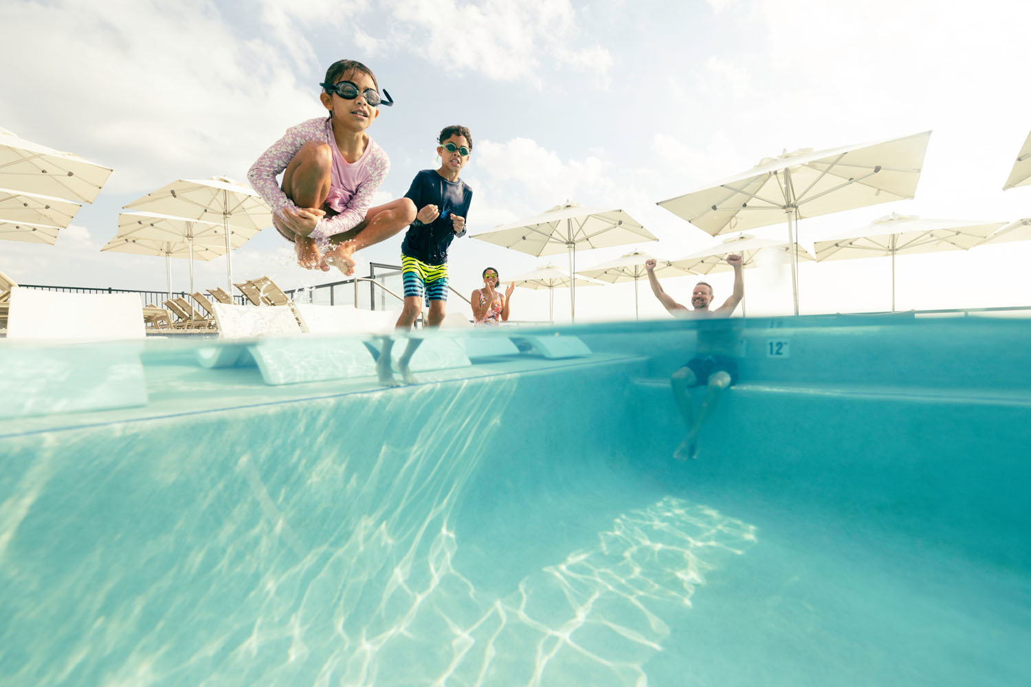 Two children jumping into an outdoor pool with their parents nearby